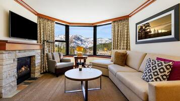A living room with a fireplace, a television, a sofa, a coffee table, and a large window with a mountain view.