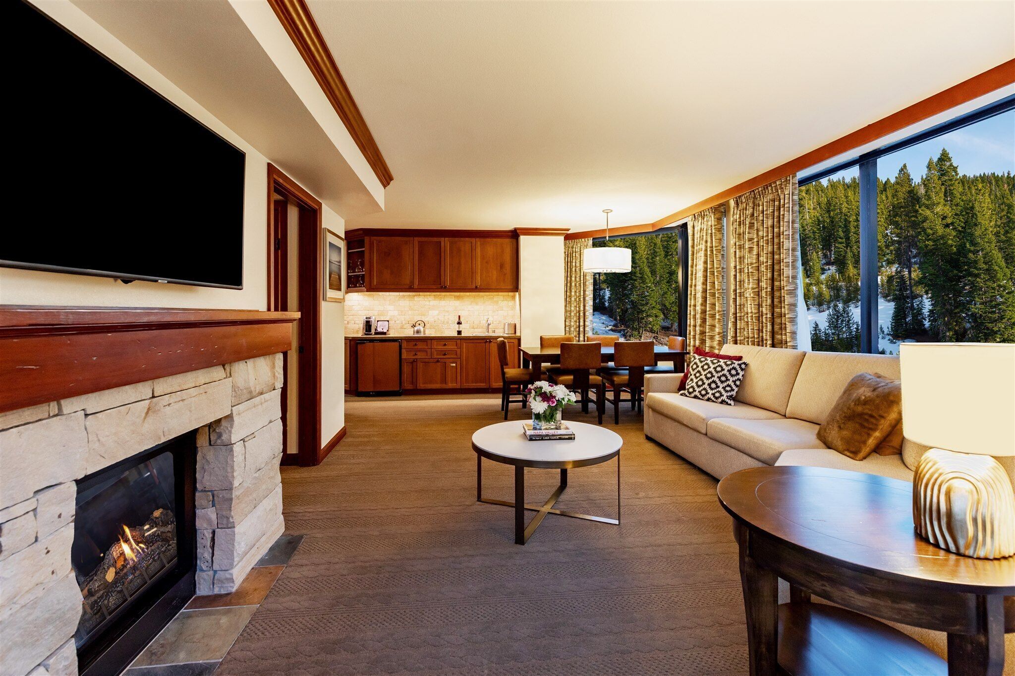 A modern living room with a stone fireplace, a flat-screen TV, a dining area with wooden cabinets, and a large window offering a view of trees.
