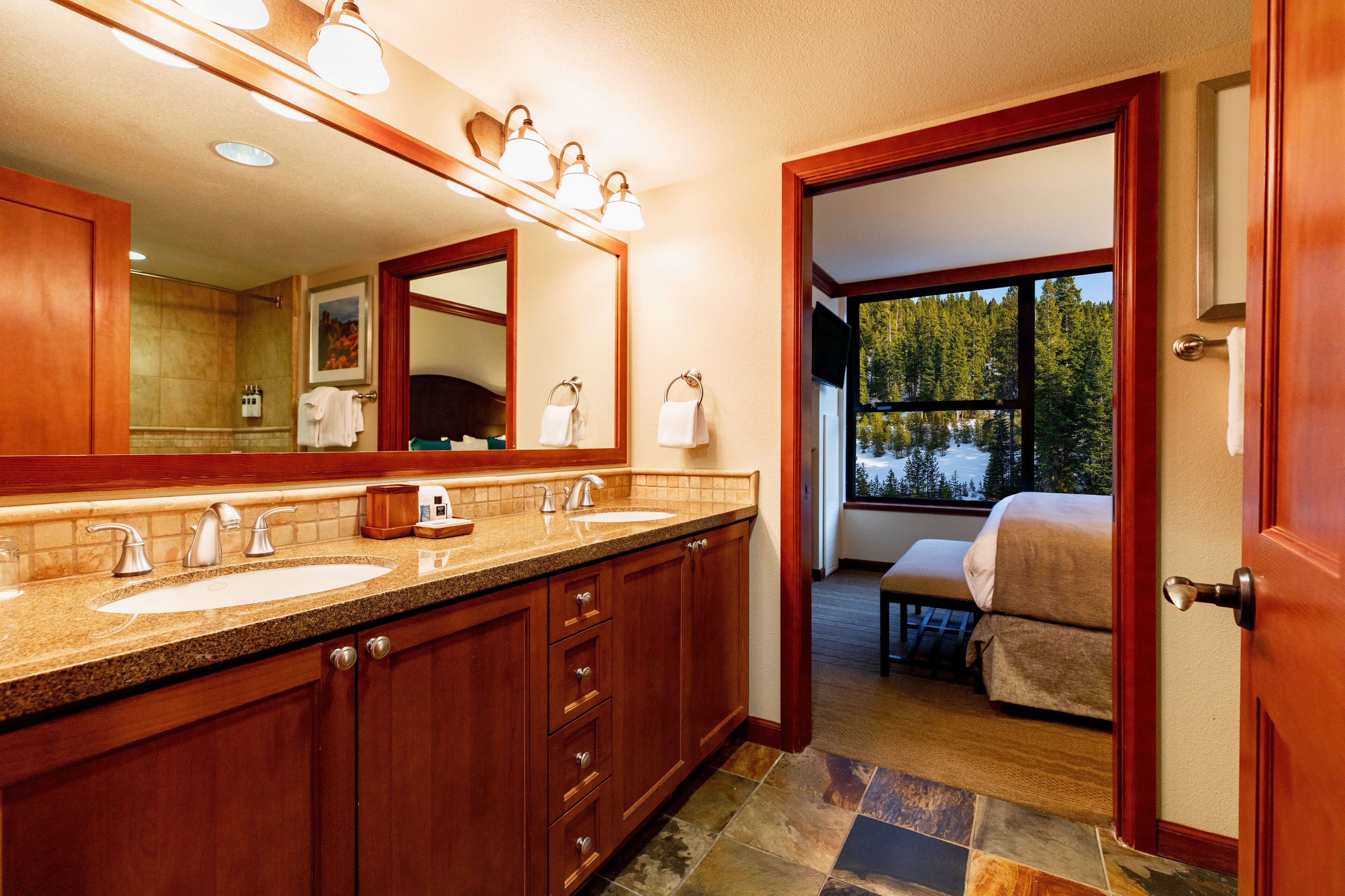 A bathroom with two sinks, a large mirror, and a view of a bedroom through an open door.