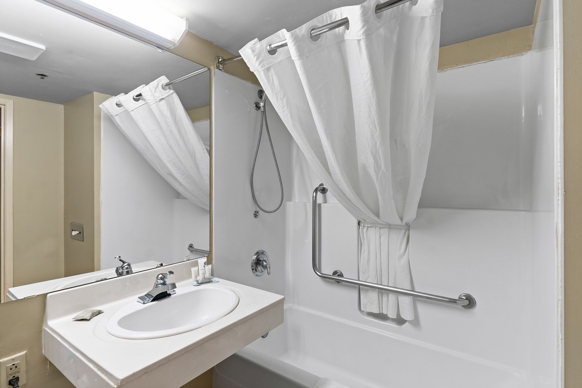 Une salle de bain avec un lavabo blanc, un miroir, une douche équipée d’une barre d’appui et un rideau de douche blanc.