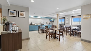 Un espace repas moderne avec des tables et des chaises en bois, une cuisine à l’arrière-plan et un téléviseur fixé au mur.