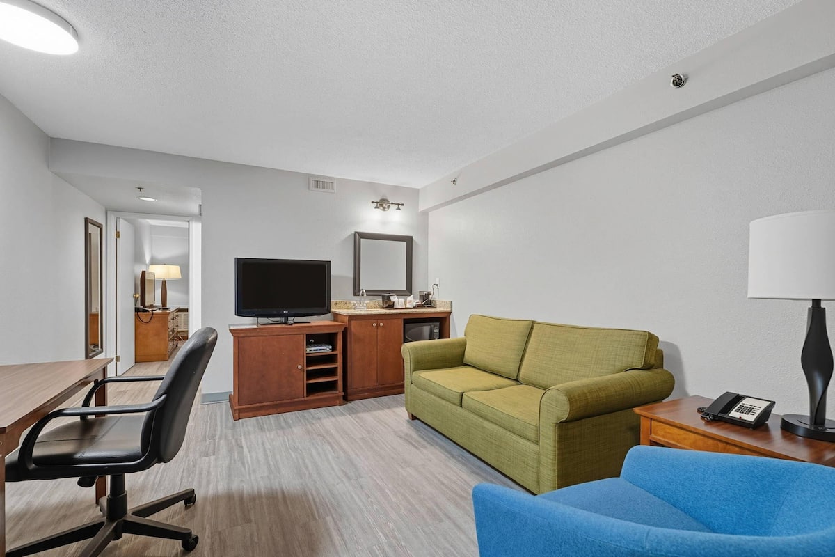 A hotel room with a wooden desk, a television, a green sofa, a blue sofa, and a telephone on a side table.
