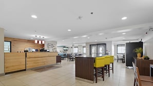 Un hall d’hôtel spacieux avec un comptoir d’accueil en bois, un espace central agrémenté de chaises jaunes et un coin repas avec des tables et des chaises.