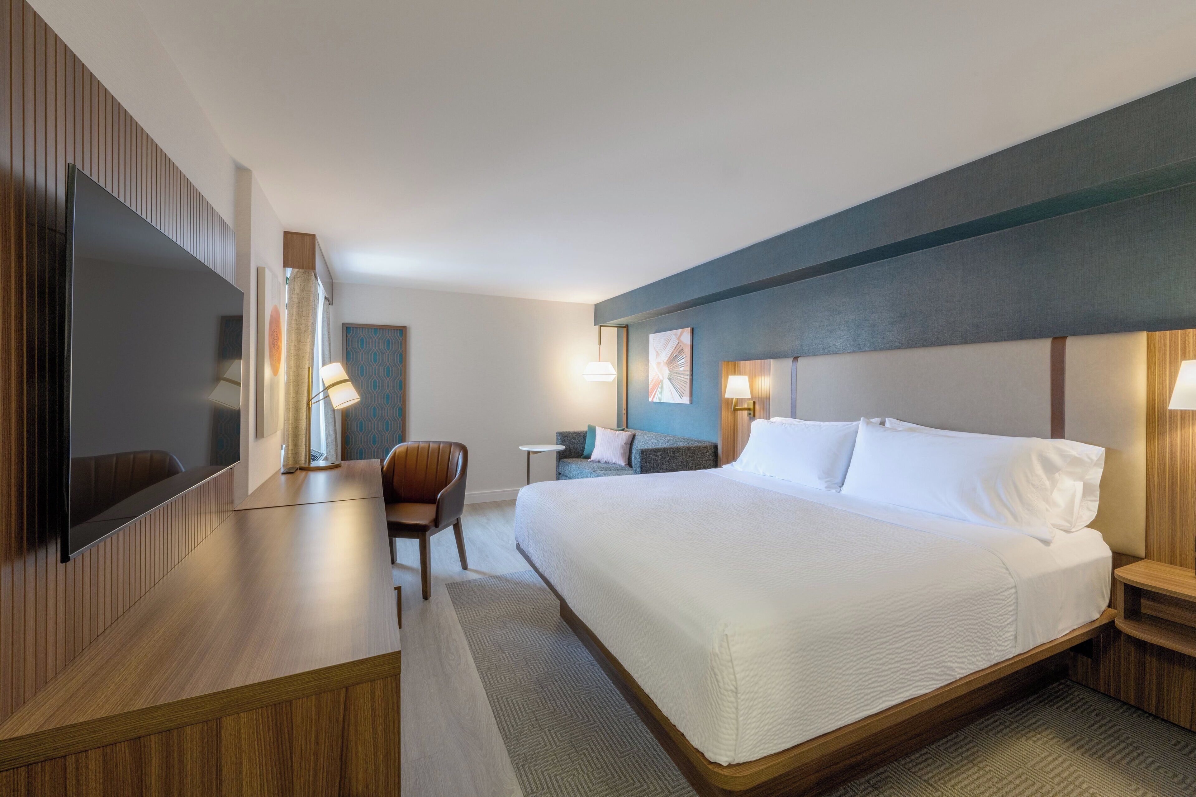A hotel room with a large bed, a desk with a flat-screen TV, a chair, and a small table.