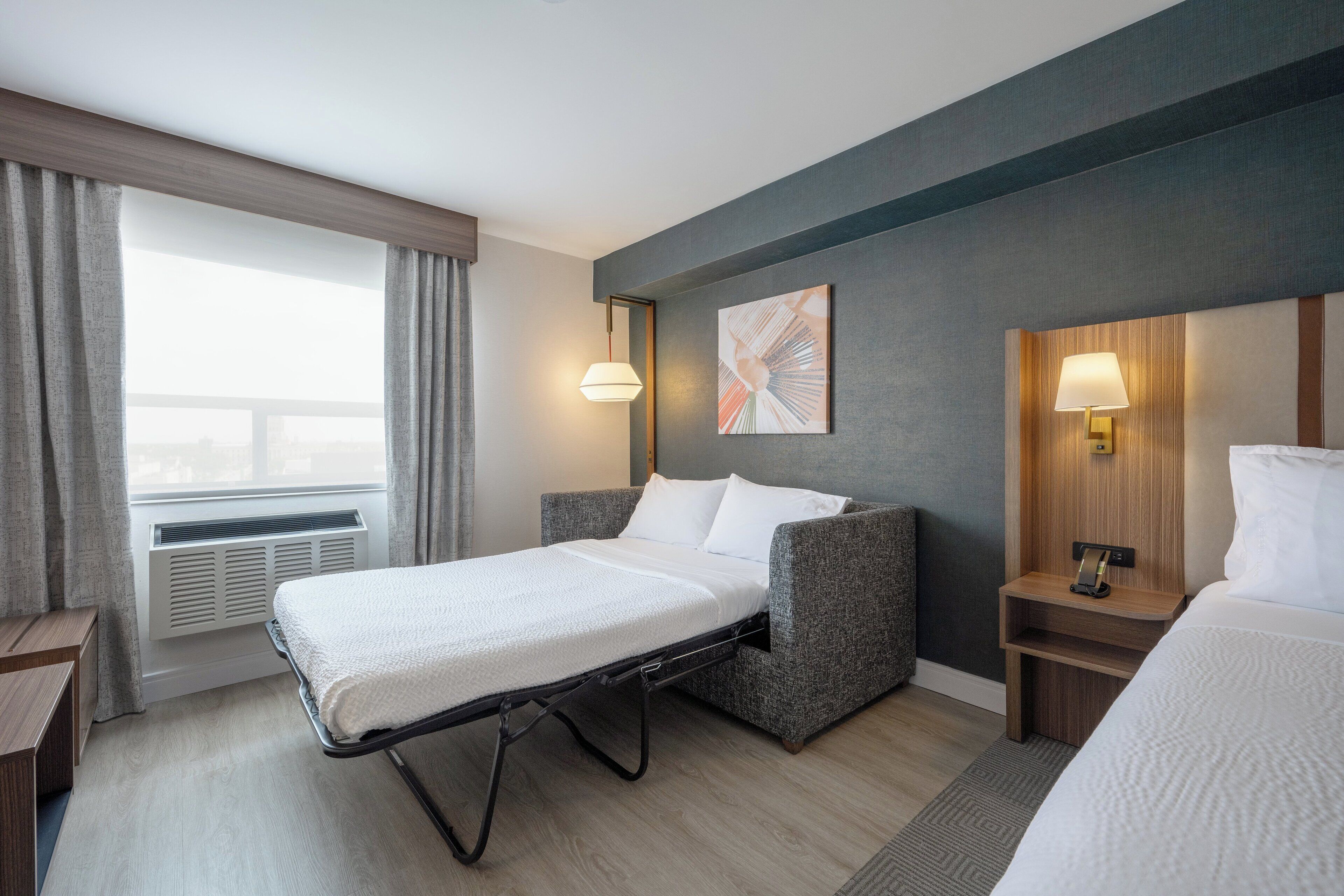 A hotel room with a bed, bedside table, lamp, artwork, and a window with curtains.