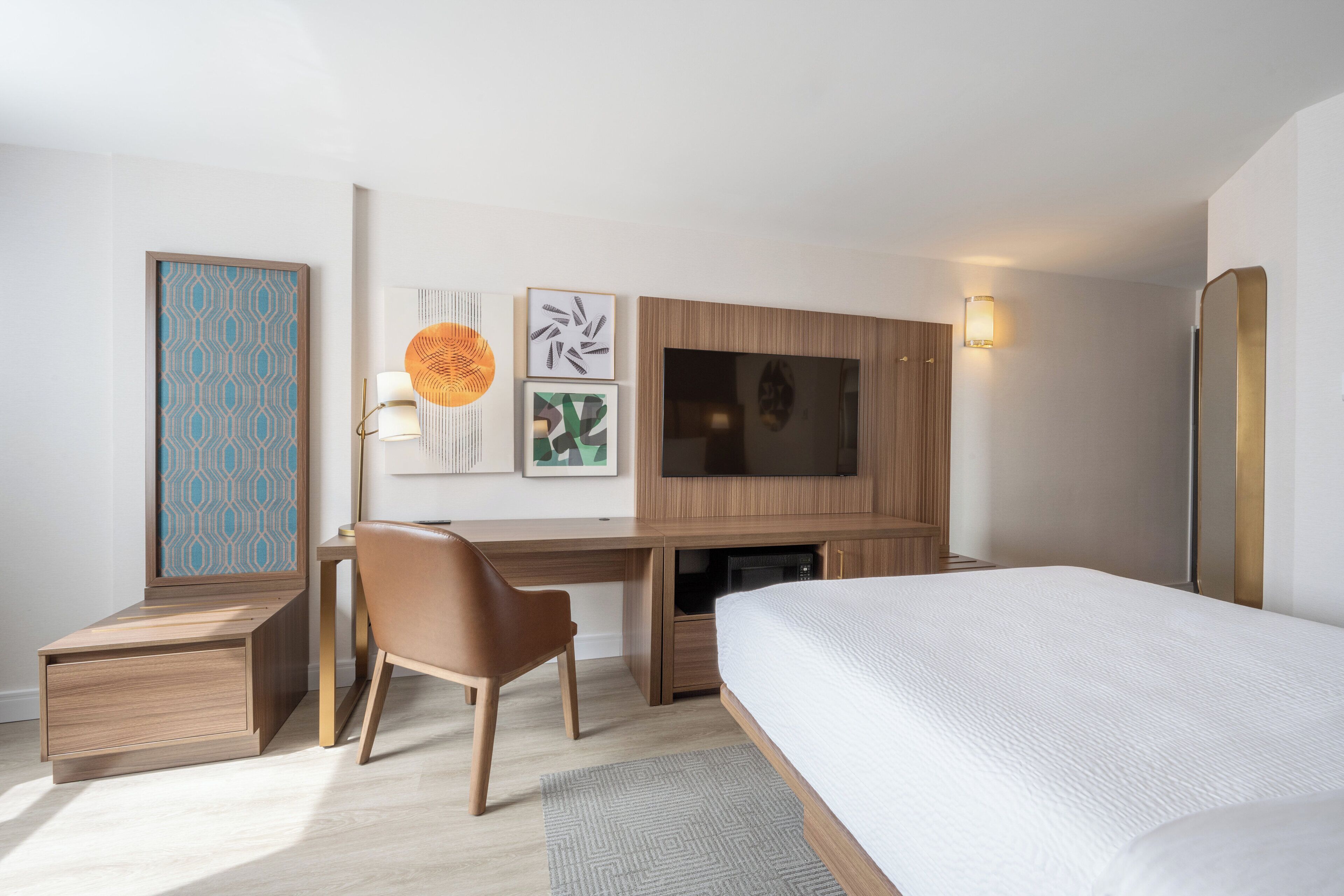 A modern hotel room with a wooden desk, a comfortable chair, a large bed, and a flat-screen TV mounted on the wall.