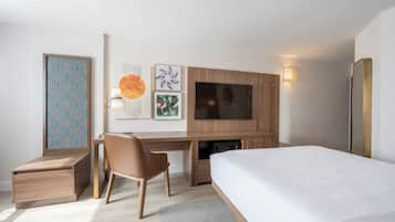 A modern hotel room with a wooden desk, a comfortable chair, a large bed, and a flat-screen TV mounted on the wall.