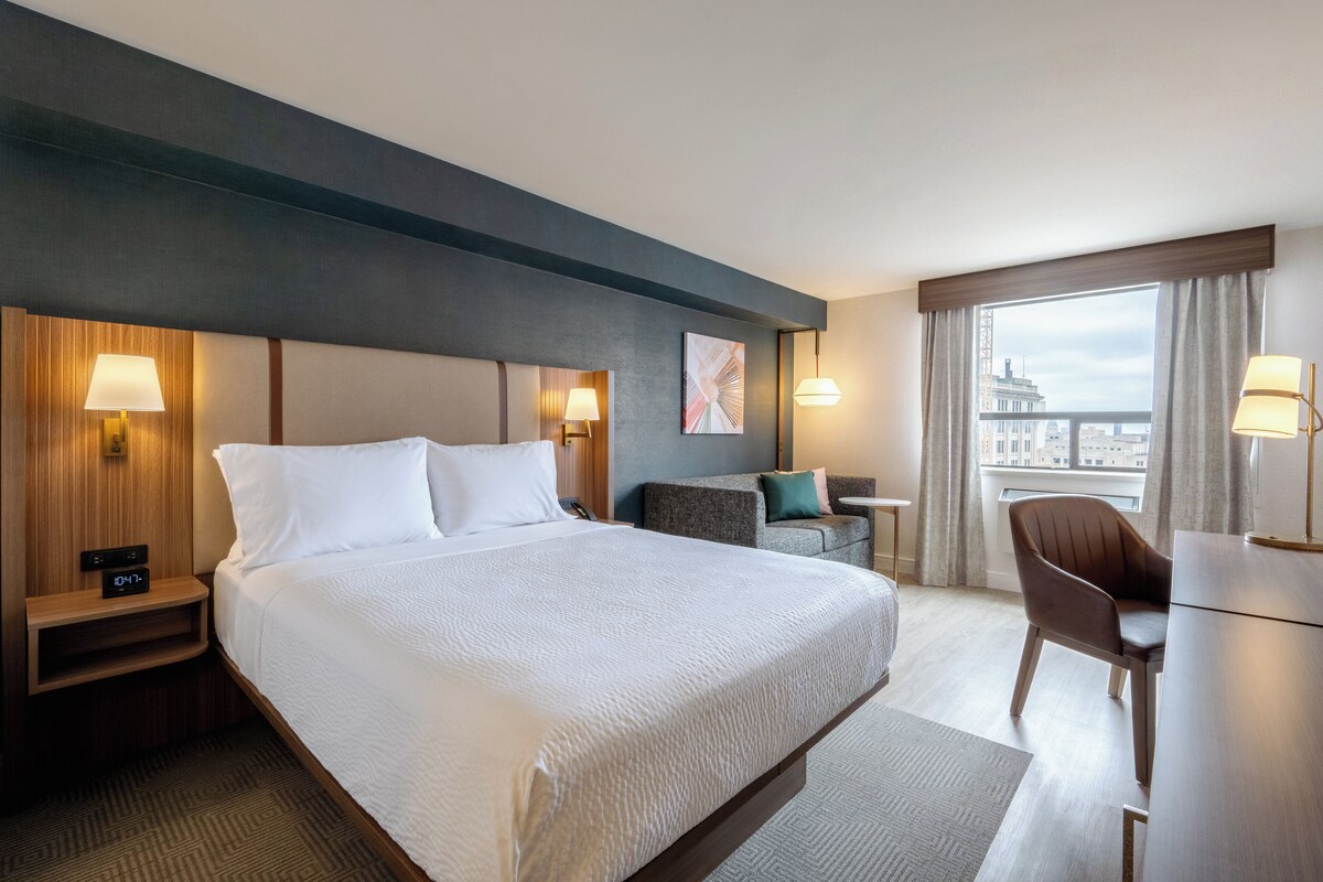 A hotel room with a large bed, bedside lamps, a desk with a chair, and a view of the city through the window.