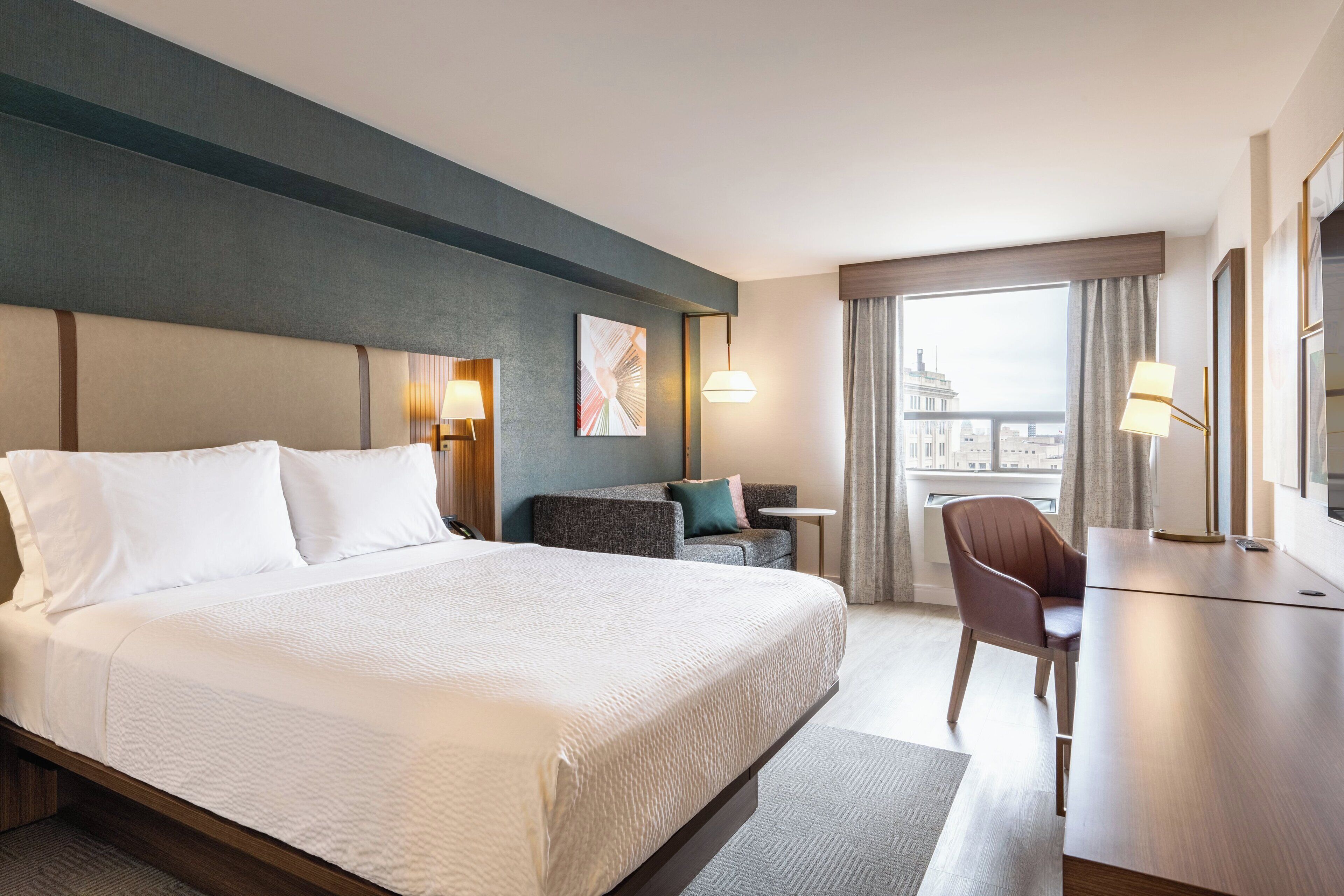 Habitación de hotel con una cama grande, un escritorio, una silla, una mesita y vistas a la ciudad a través del ventanal.
