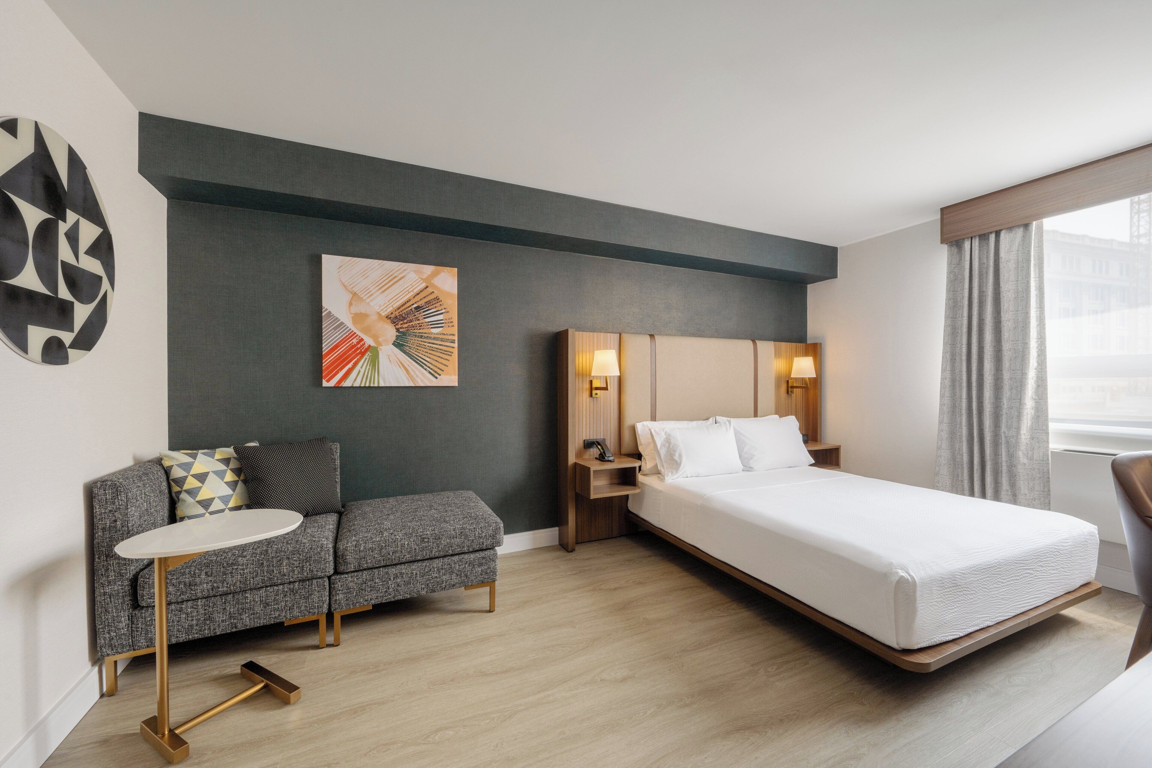 A modern hotel room with a bed, a sofa, a small table, and a wall art piece.