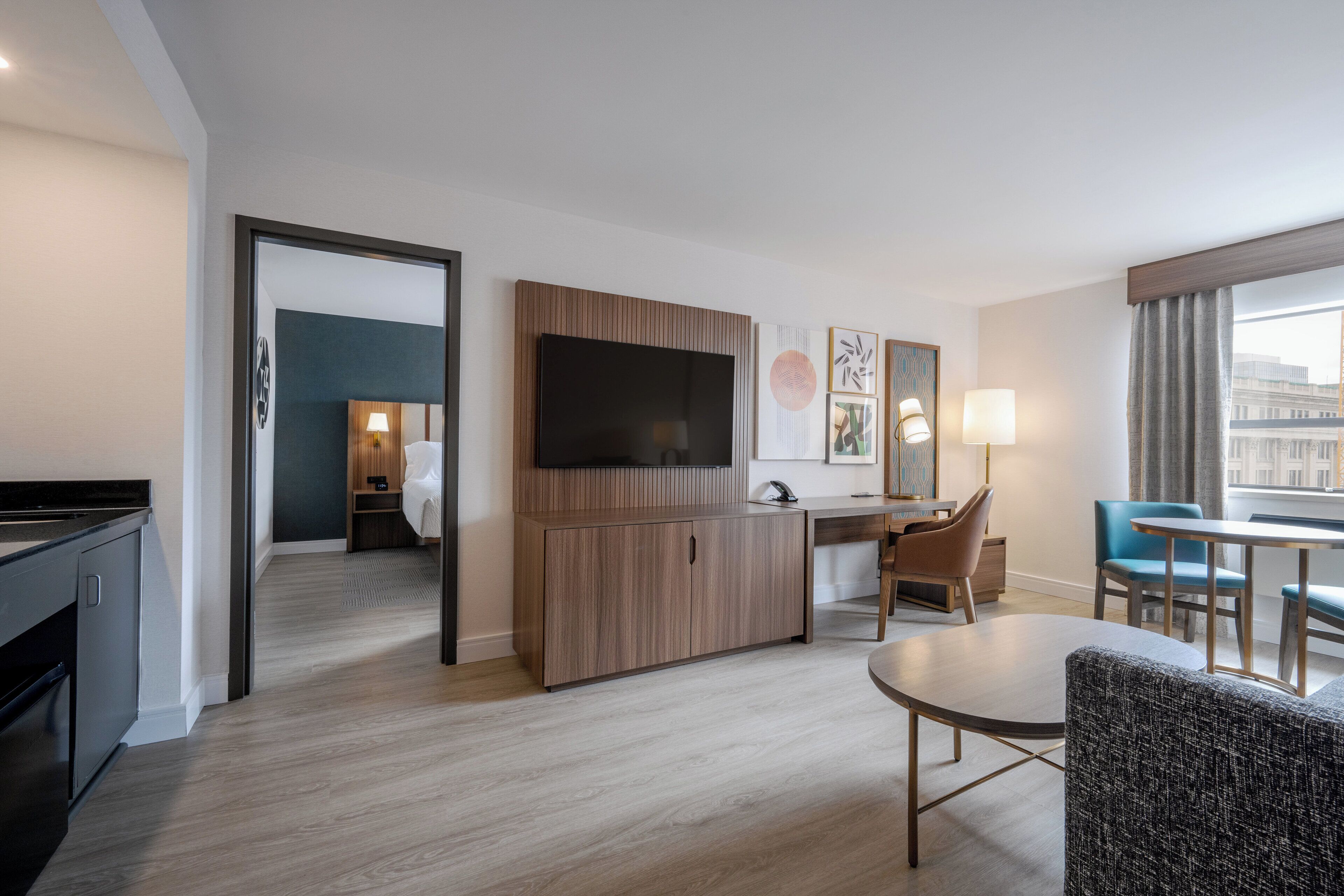 Habitación de hotel moderna con televisión de pantalla plana, un armario de madera, zona de comedor con mesa y sillas, y vista a la ciudad a través de amplios ventanales.