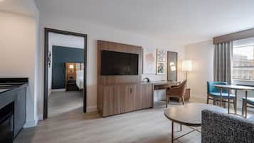 Habitación de hotel moderna con televisión de pantalla plana, un armario de madera, zona de comedor con mesa y sillas, y vista a la ciudad a través de amplios ventanales.