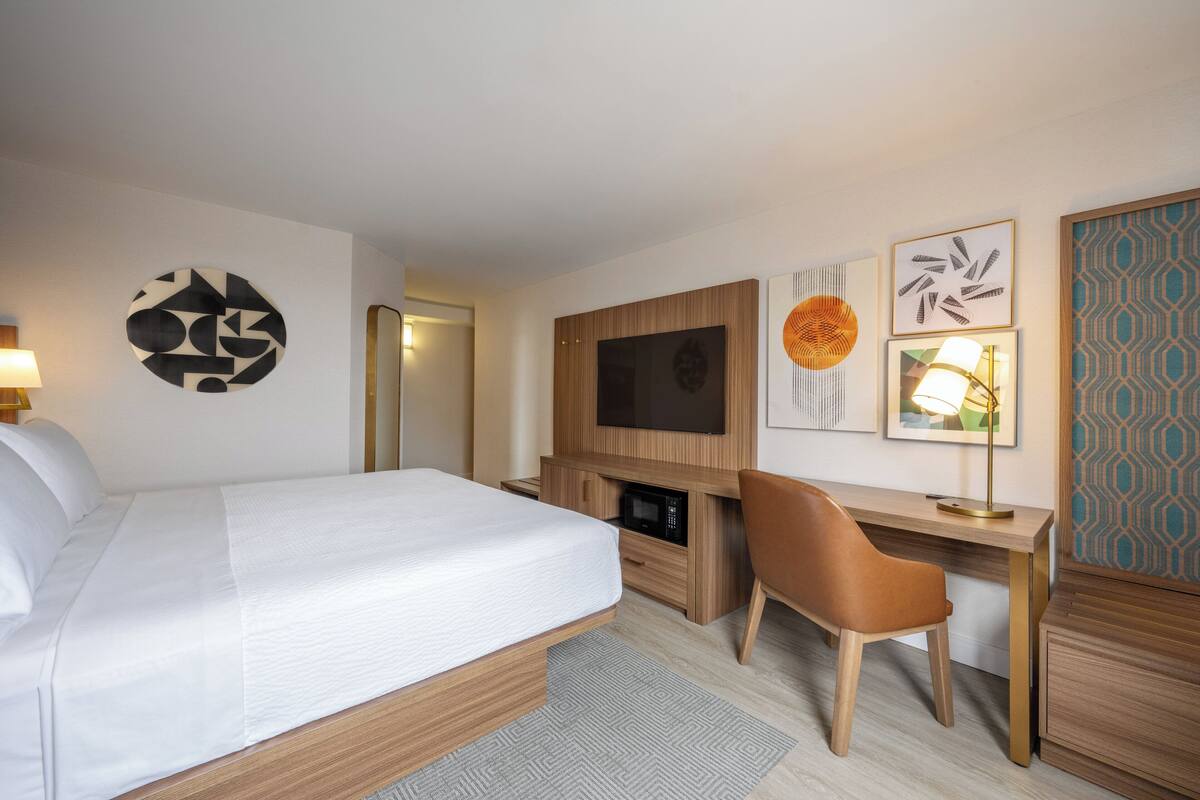 A hotel room with a bed, a desk, a chair, a TV, and artwork on the walls.