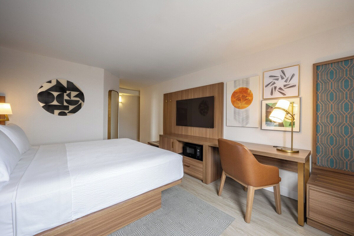 A hotel room with a bed, a desk, a chair, a TV, and artwork on the walls.