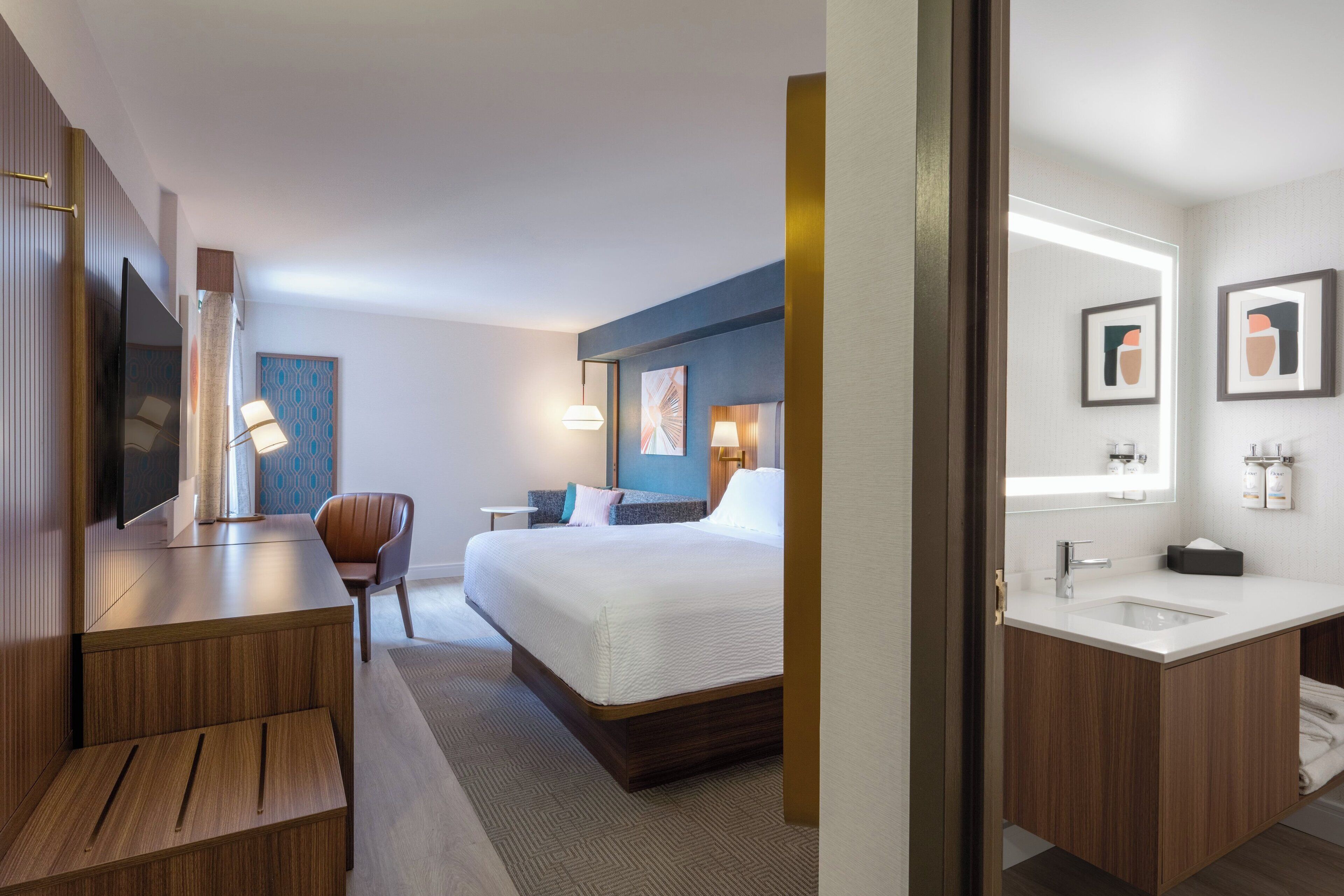 Habitación de hotel con cama, mesita de noche, silla y baño con lavabo y espejo.