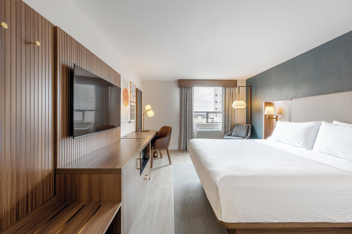 A hotel room with a large bed, a desk with a flat-screen TV, a chair, and a window with curtains.