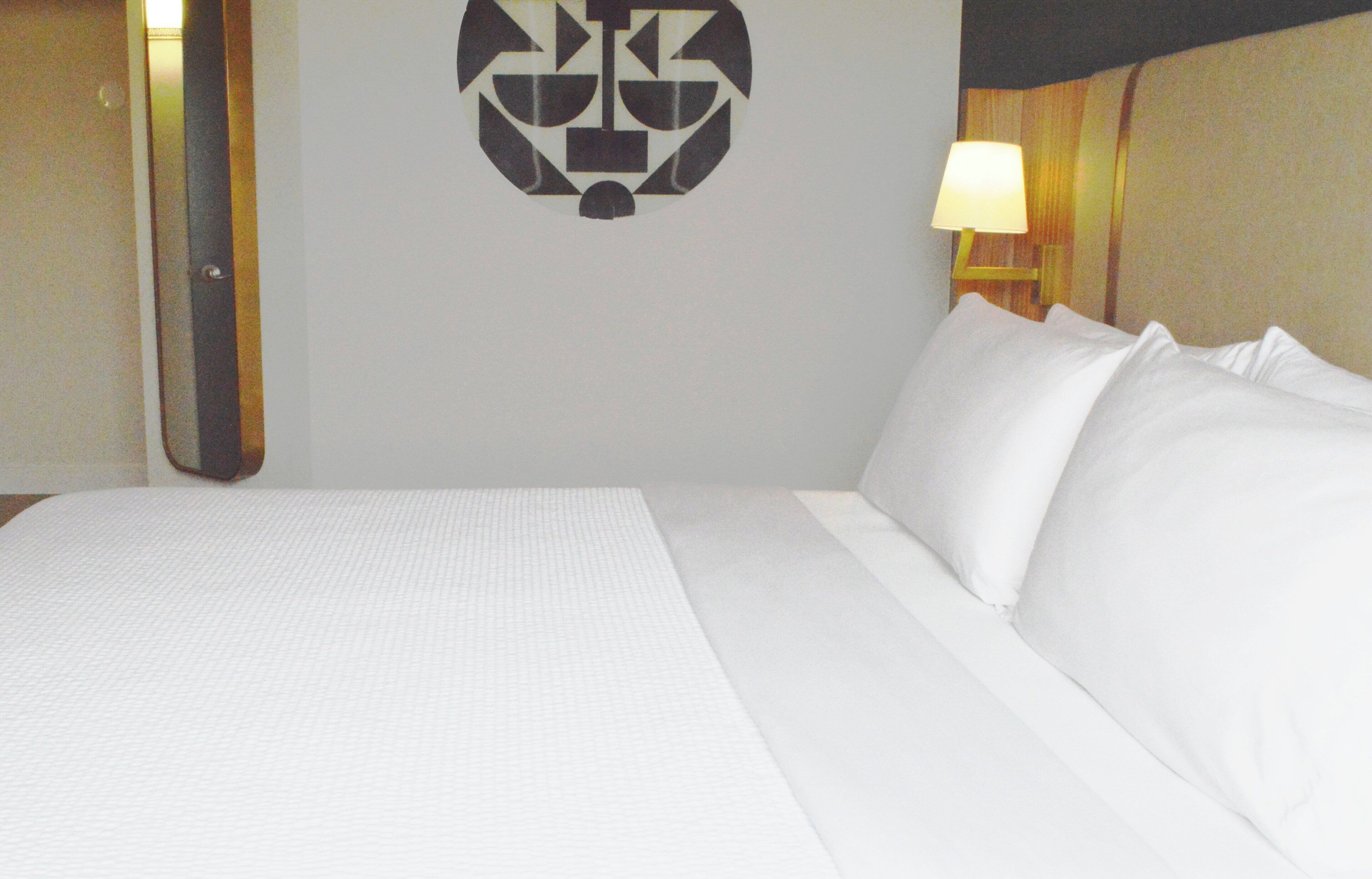 A hotel room with a bed, pillows, a wall-mounted logo, and a nightstand with a lamp.