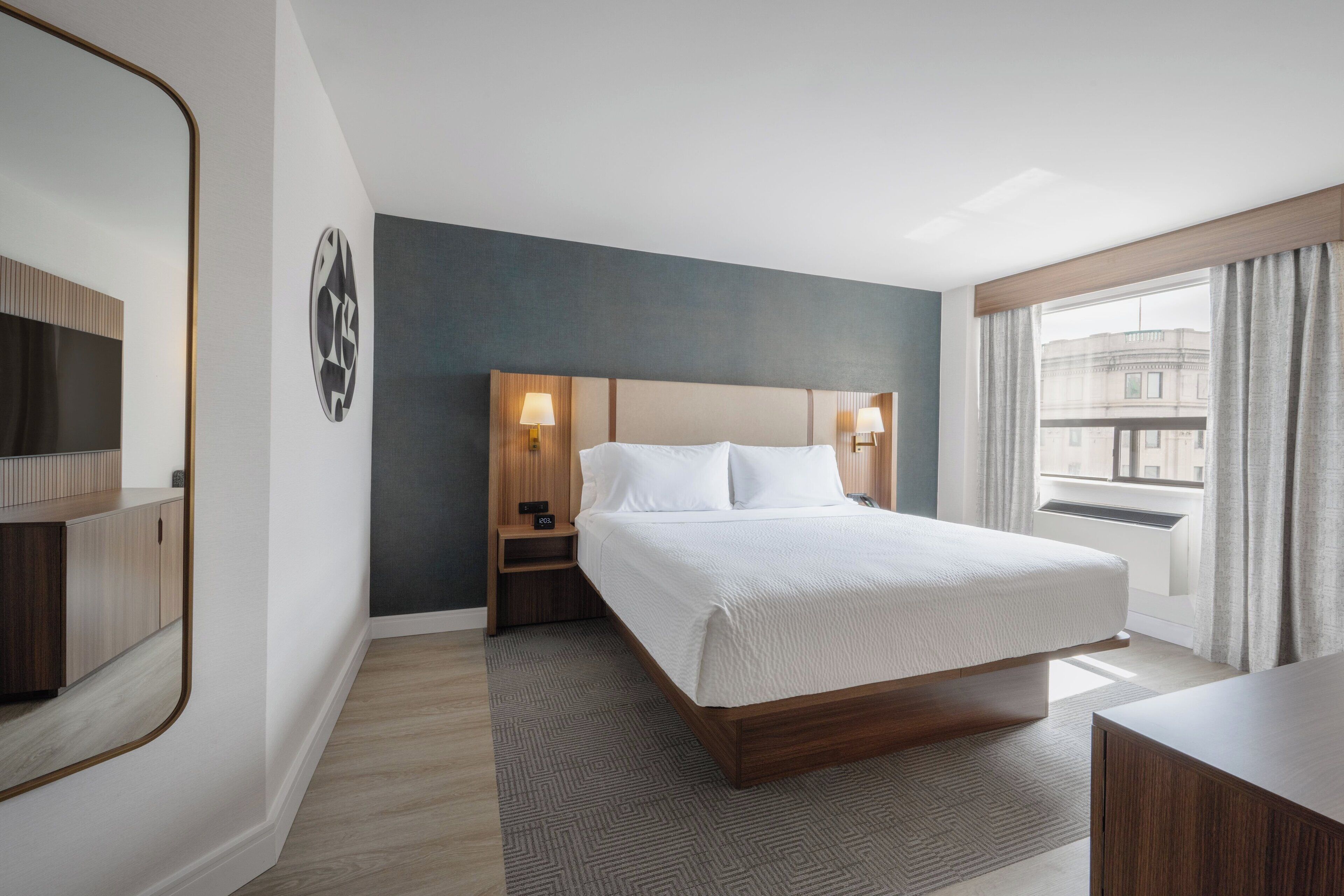 Habitación de hotel con una cama grande, lámparas de noche, mesita de noche, un espejo y vista a la ciudad por la ventana.