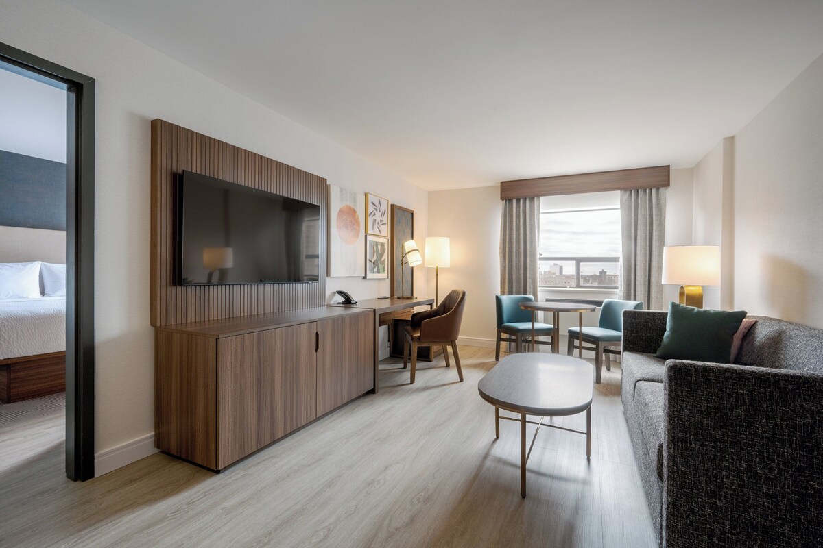 A modern hotel room with a wooden desk, a television, a sofa, and a dining area.