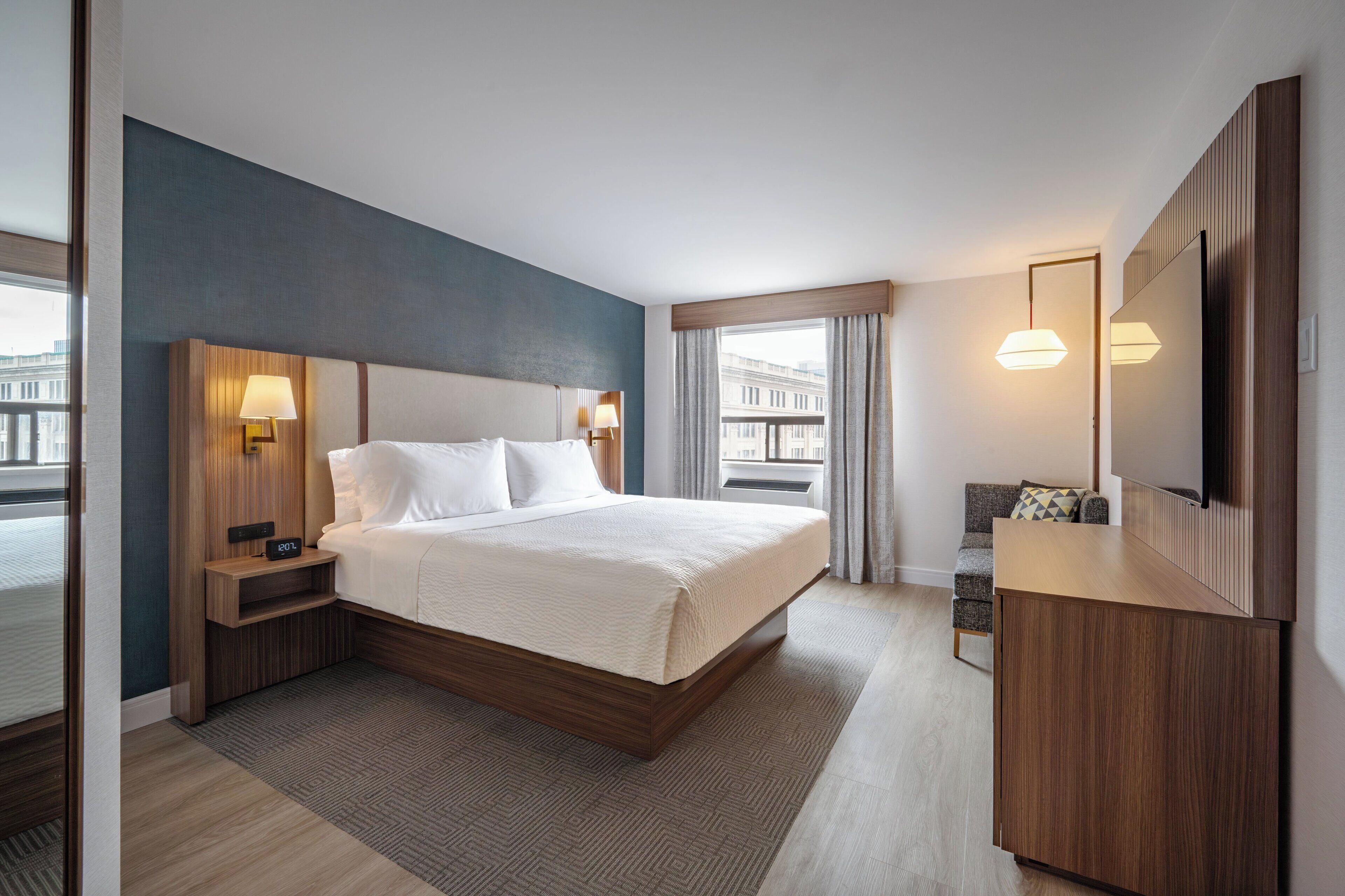 A modern hotel room with a large bed, bedside tables with lamps, a flat-screen TV, and a view of the city through the window.