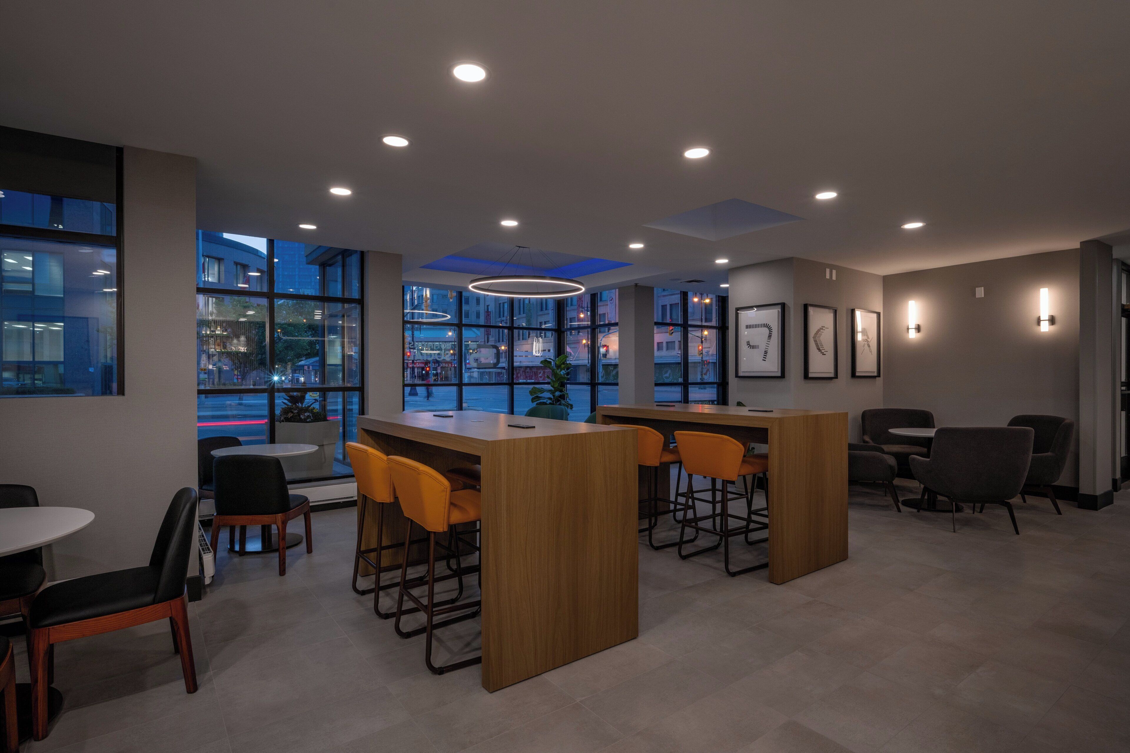 A modern bar area with wooden counters and orange chairs, large windows with city views, and recessed ceiling lighting.