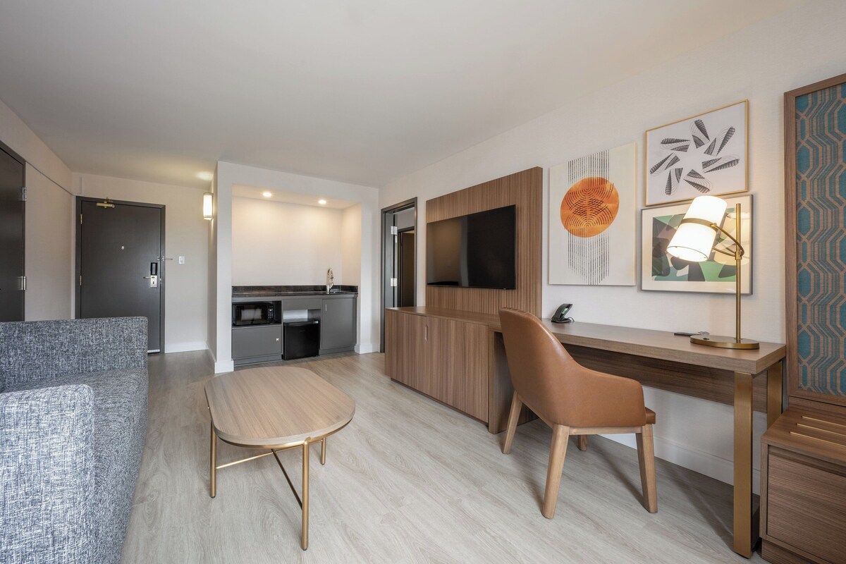 A modern hotel room with a sofa, a dining table, a desk with a lamp, a TV, and a kitchen area.