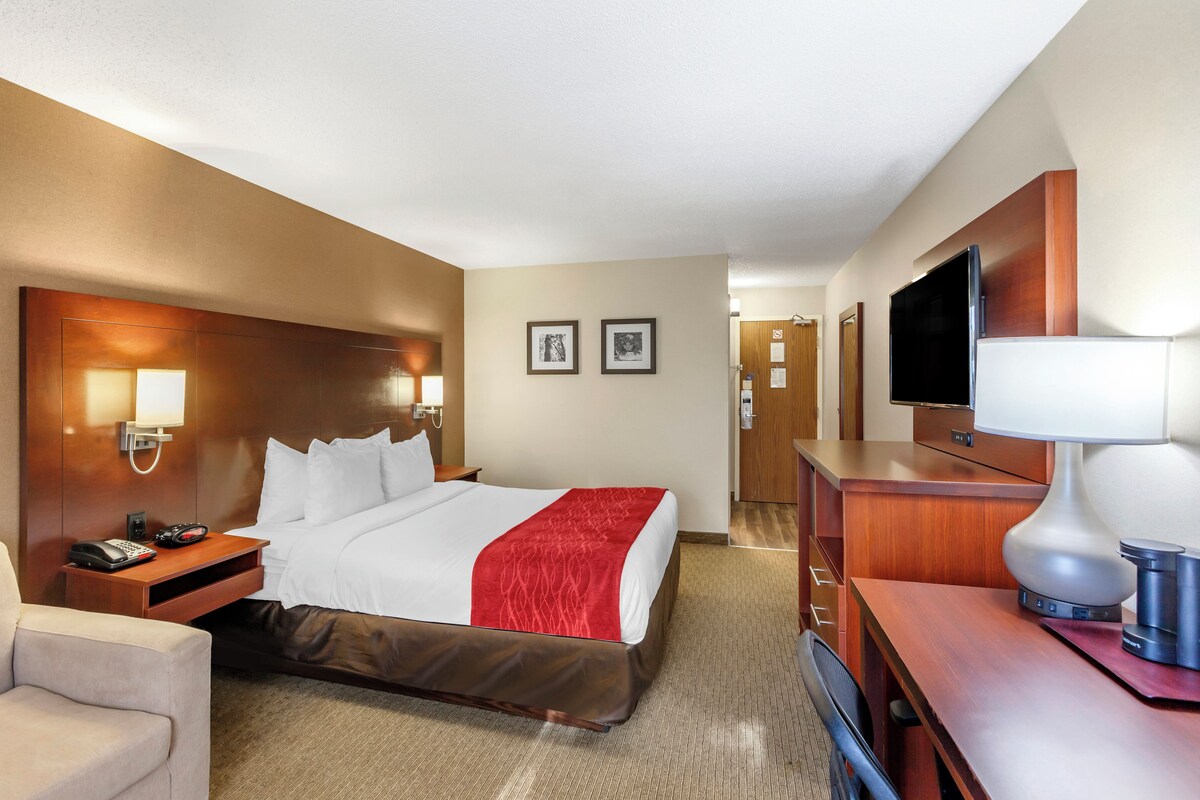 A hotel room with a bed, bedside table, lamp, TV, and a desk with a chair.