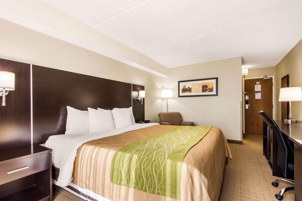 A hotel room with a bed, a desk, a chair, a lamp, and a door.