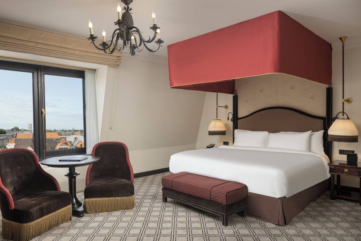 Une chambre d’hôtel avec un grand lit, deux fauteuils, une petite table, un lit à baldaquin rouge, un lustre et une vue sur la ville.