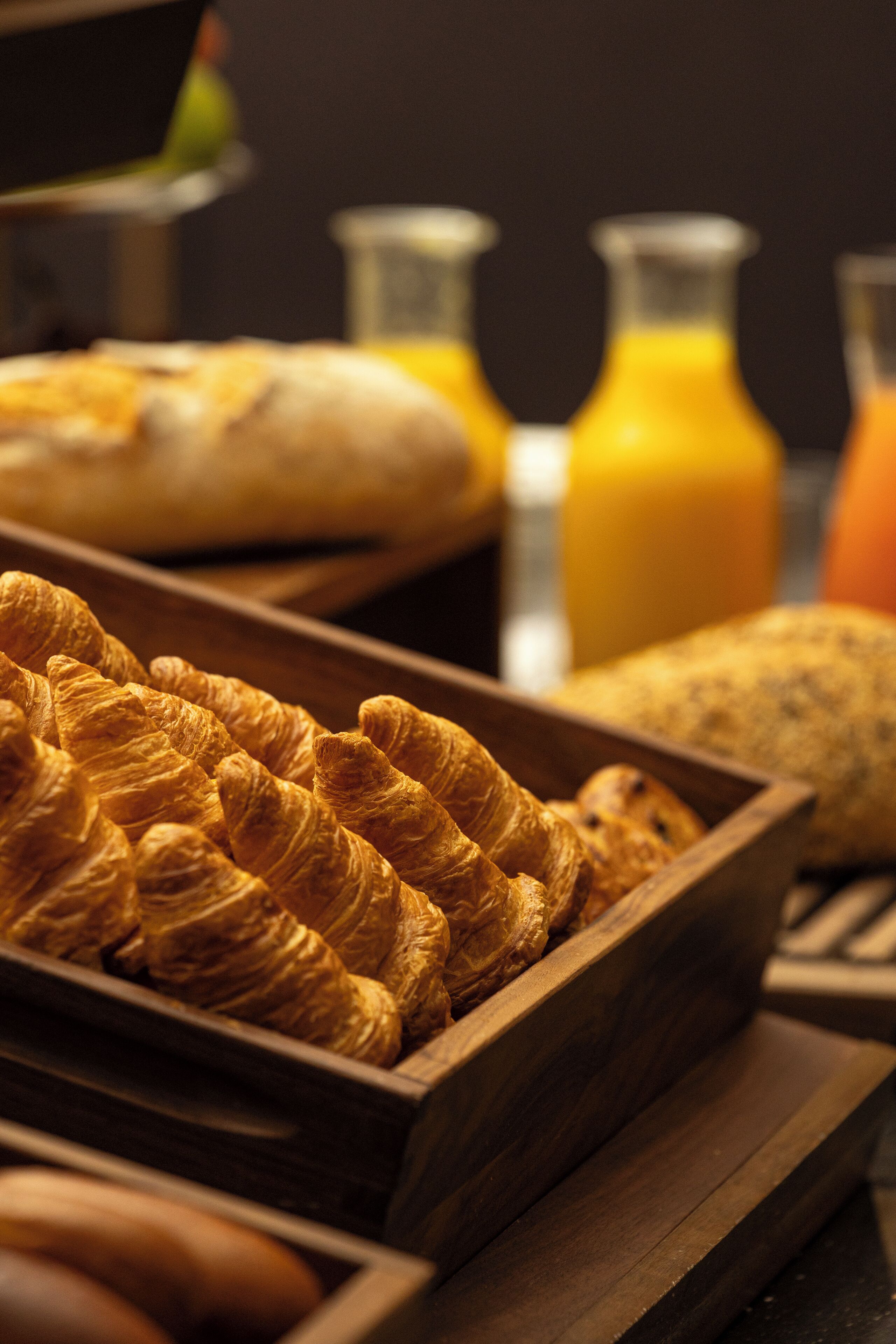 A wooden tray filled with croissants and a loaf of bread, with a glass bottle of orange juice in the background.