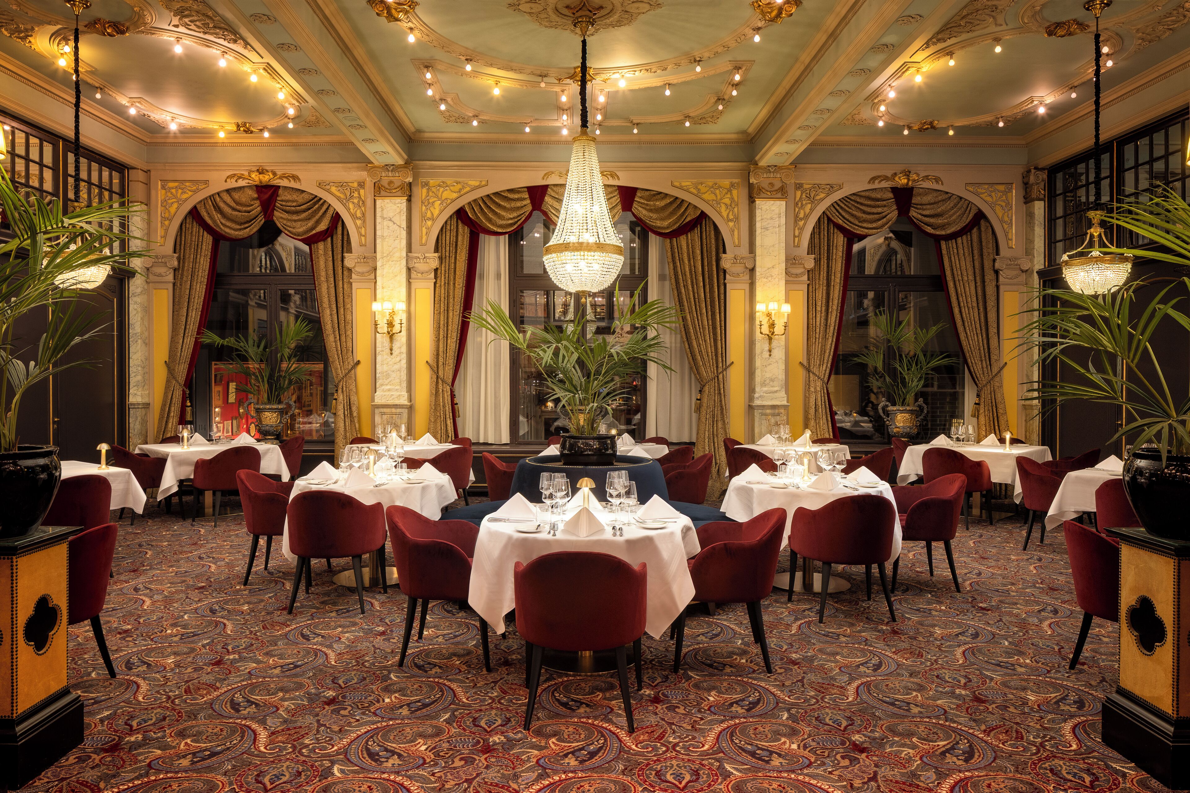 Uma sala de jantar formal, com mesas postas para servir, lustres ornamentados e arquitetura clássica.