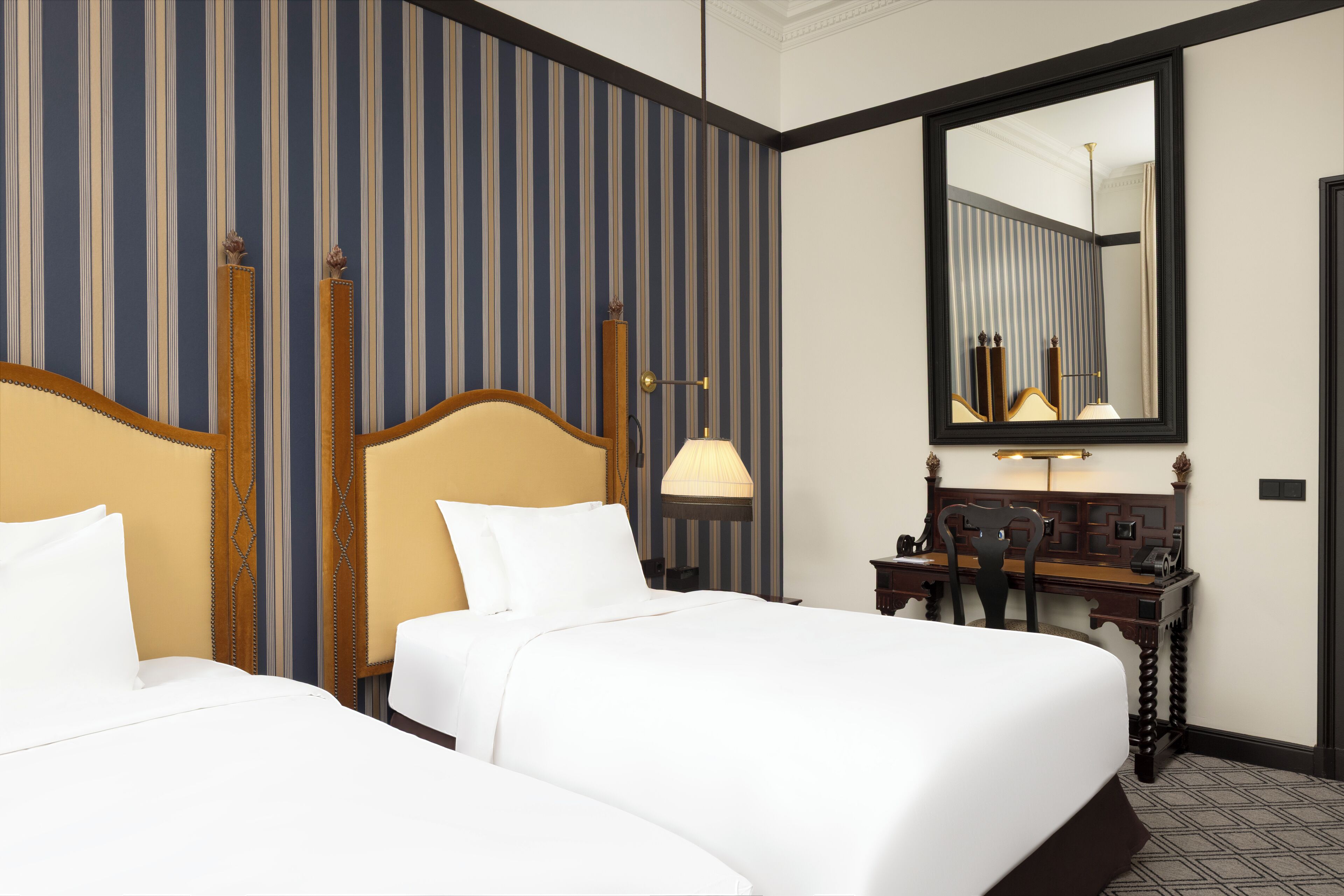 A hotel room with two beds, a desk, a chair, a lamp, and a large mirror.