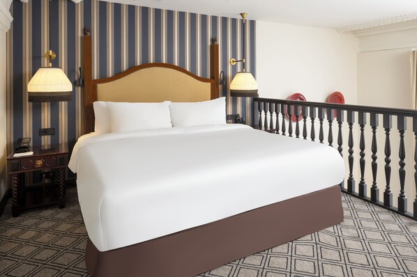 A hotel room with a large bed, striped wallpaper, two bedside lamps, a wooden nightstand, and a balcony with a railing.