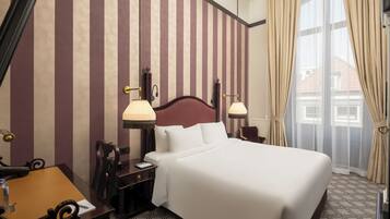 A hotel room with a large bed, two bedside lamps, a wooden nightstand, a desk with a chair, and a window with curtains.
