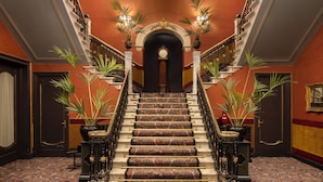 Una imponente escalera con barandillas ornamentadas y plantas en macetas, que conduce a un vano oscuro.
