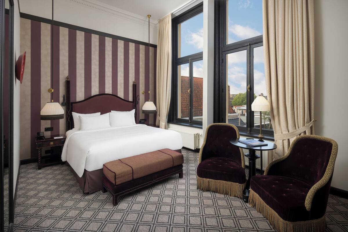 Une chambre d’hôtel avec un grand lit, une table de chevet, une chaise, une petite table et une fenêtre avec des rideaux.
