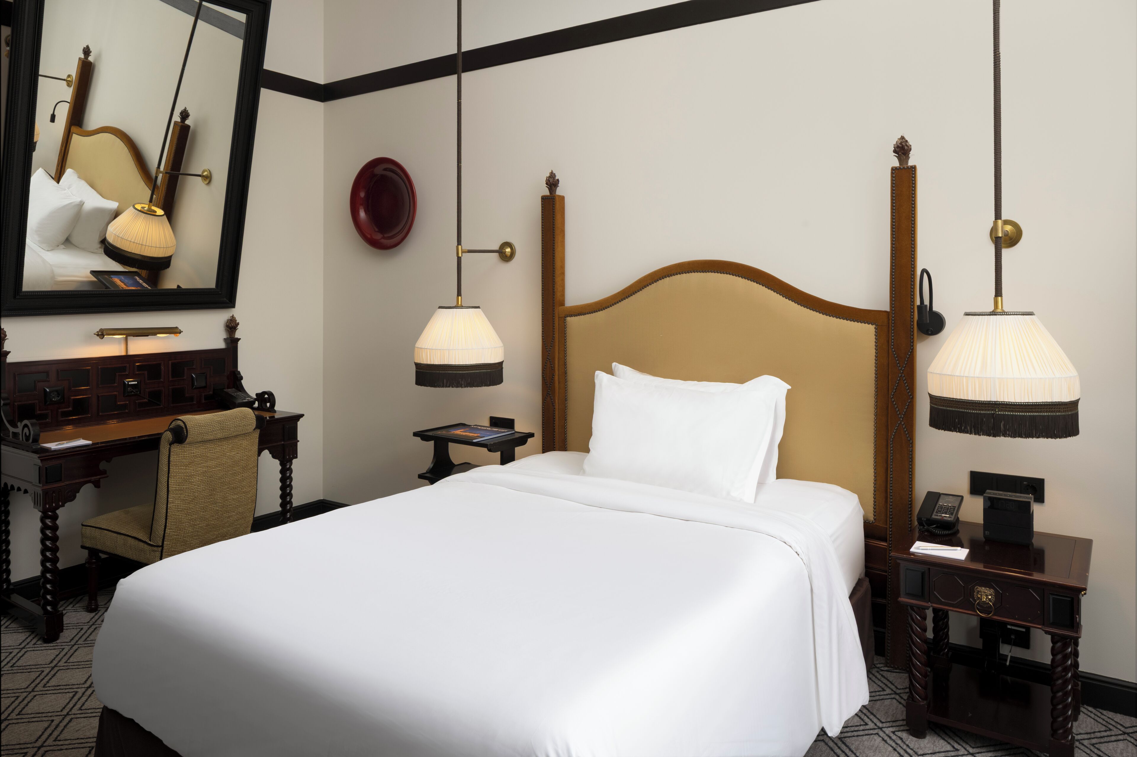 Quarto de hotel com uma cama grande, uma escrivaninha de madeira, uma cadeira, uma mesa de cabeceira com abajur e um espelho na parede.
