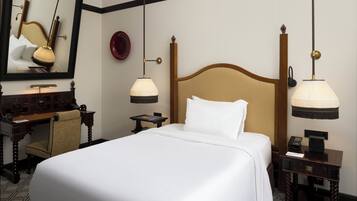 Quarto de hotel com uma cama grande, uma escrivaninha de madeira, uma cadeira, uma mesa de cabeceira com abajur e um espelho na parede.