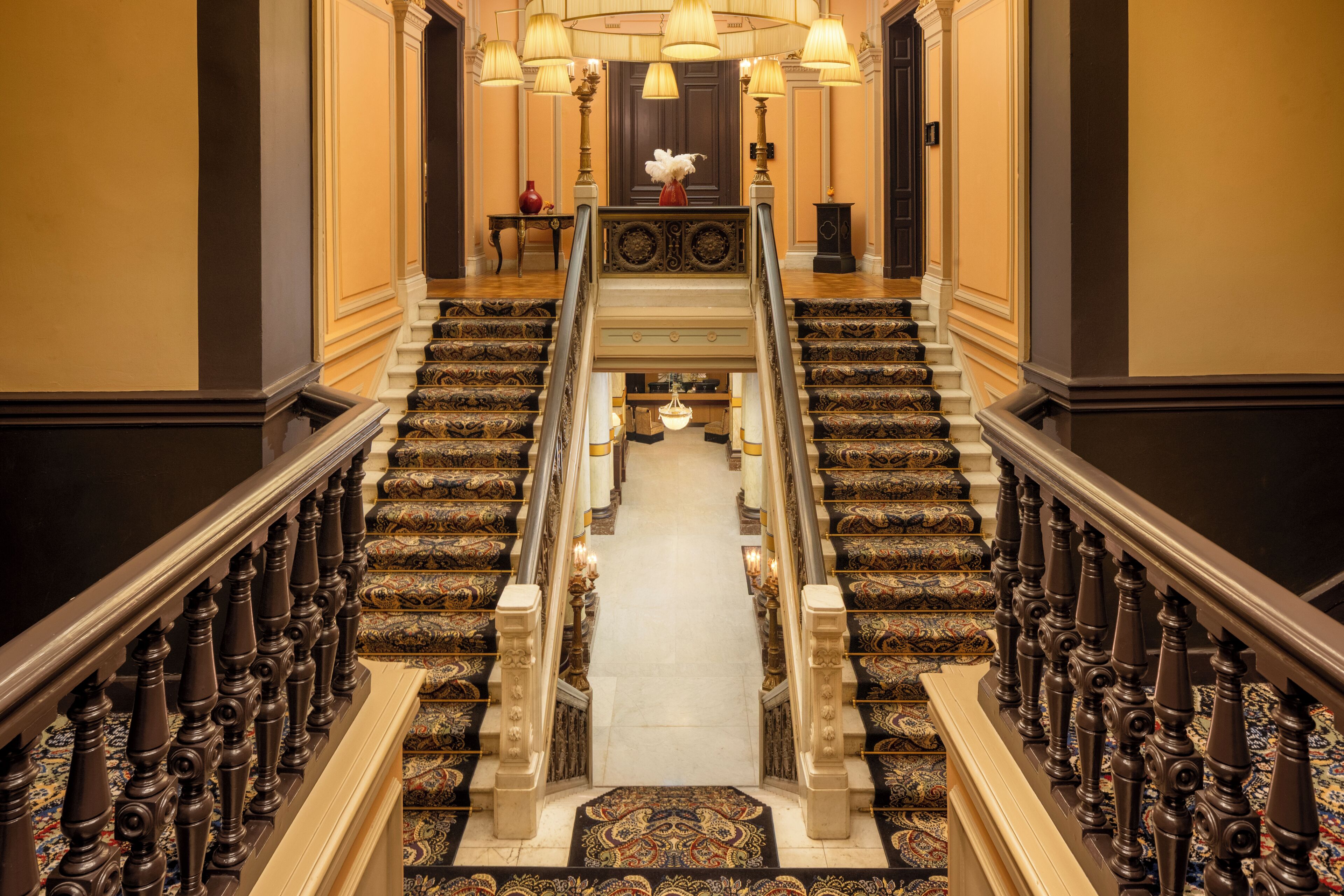 A grand staircase with ornate railings and carpeted steps leading to a landing with a chandelier.