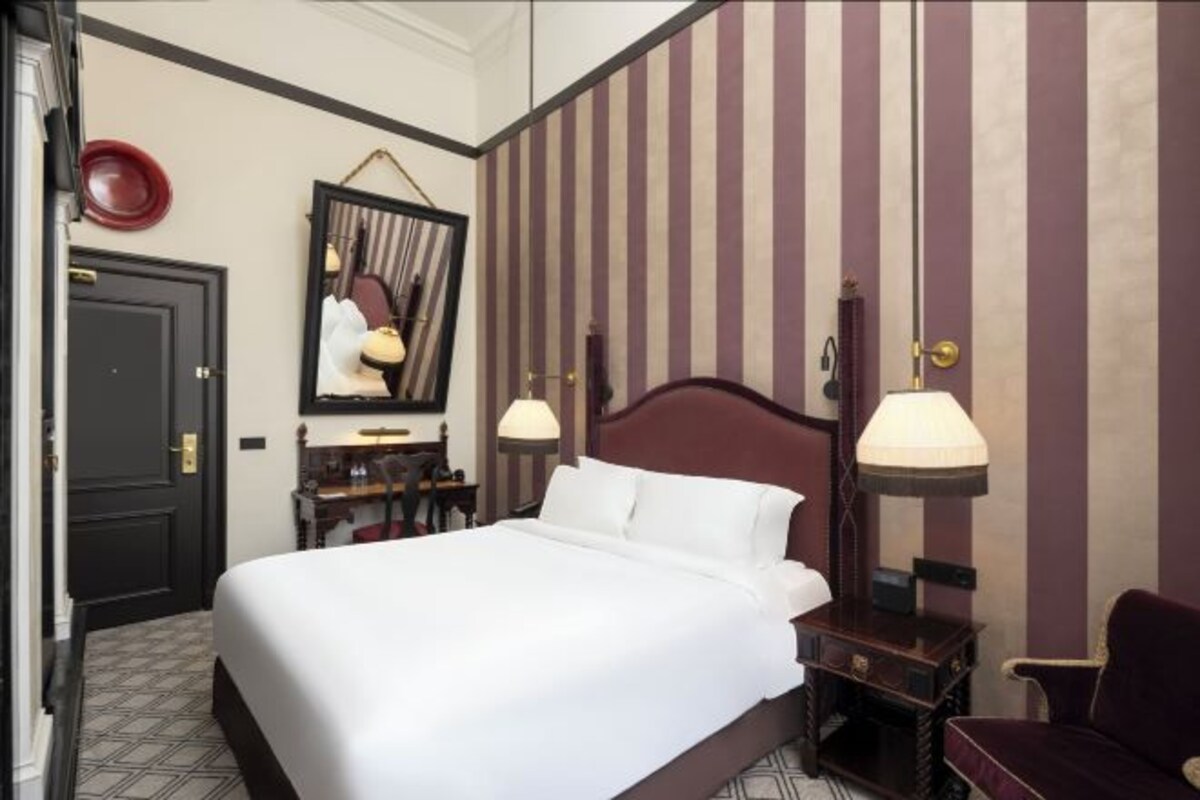 A hotel room with a bed, bedside tables, a mirror, a chair, and striped wallpaper.