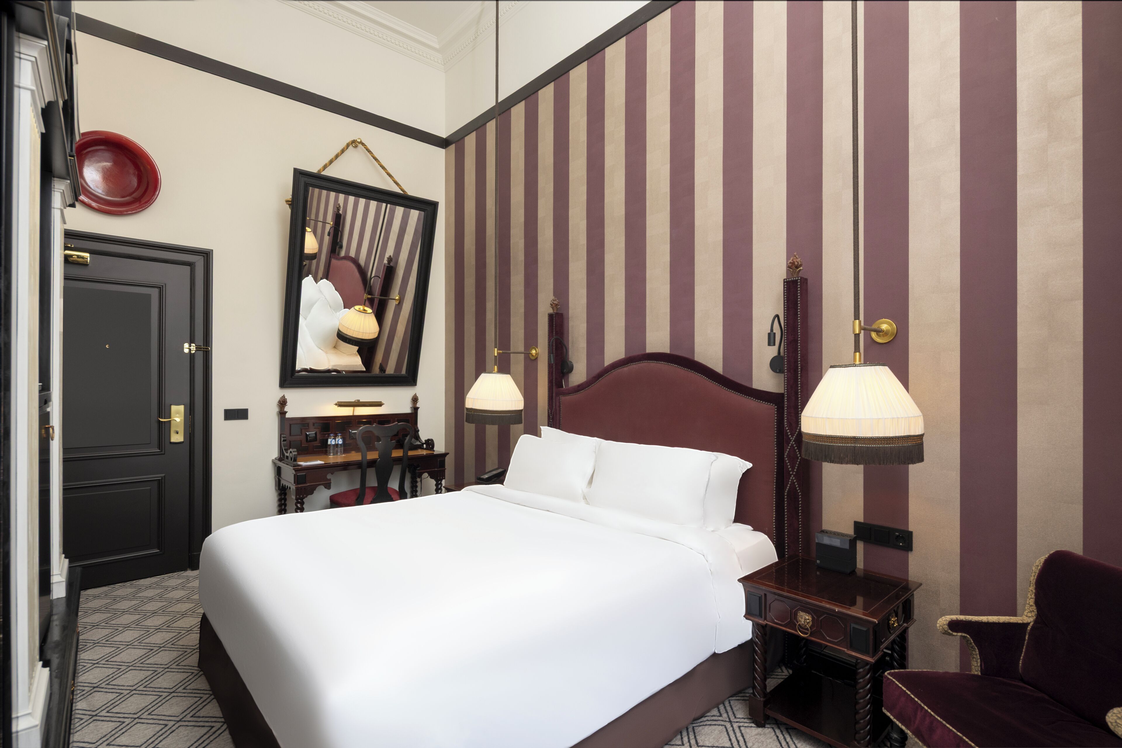 A hotel room with a large bed, a desk with a chair, a mirror, and two wall-mounted lamps.