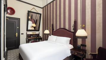 A hotel room with a large bed, a desk with a chair, a mirror, and two wall-mounted lamps.
