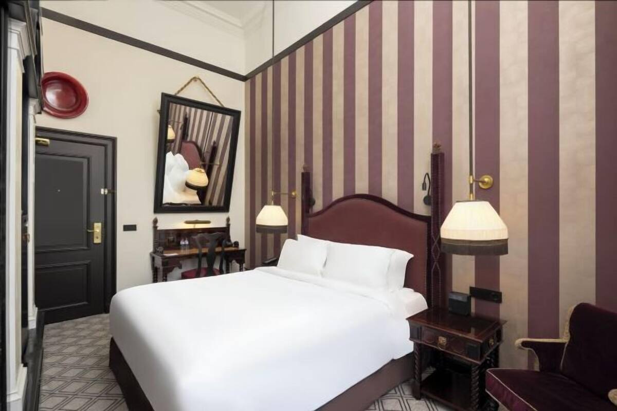 A hotel room with a bed, bedside tables, a mirror, a chair, and striped wallpaper.