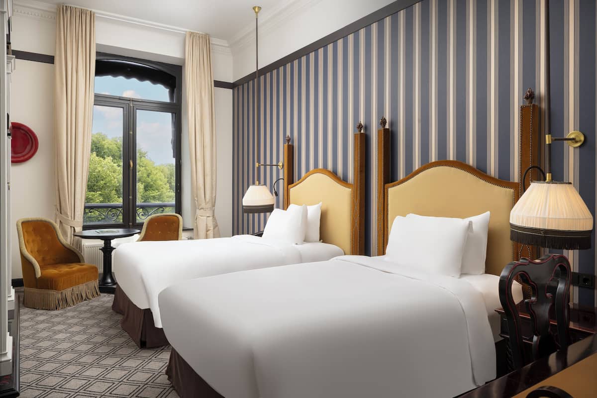 A hotel room with two beds, a window with curtains, a table, and a chair.