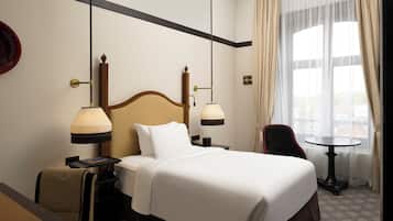 Ett hotellrum med en stor säng, två sänglampor, ett litet bord, en stol och utsikt över omgivningarna genom stora fönster.