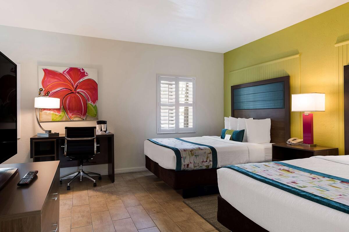 A hotel room with a desk, two beds, a large window, and a painting on the wall.