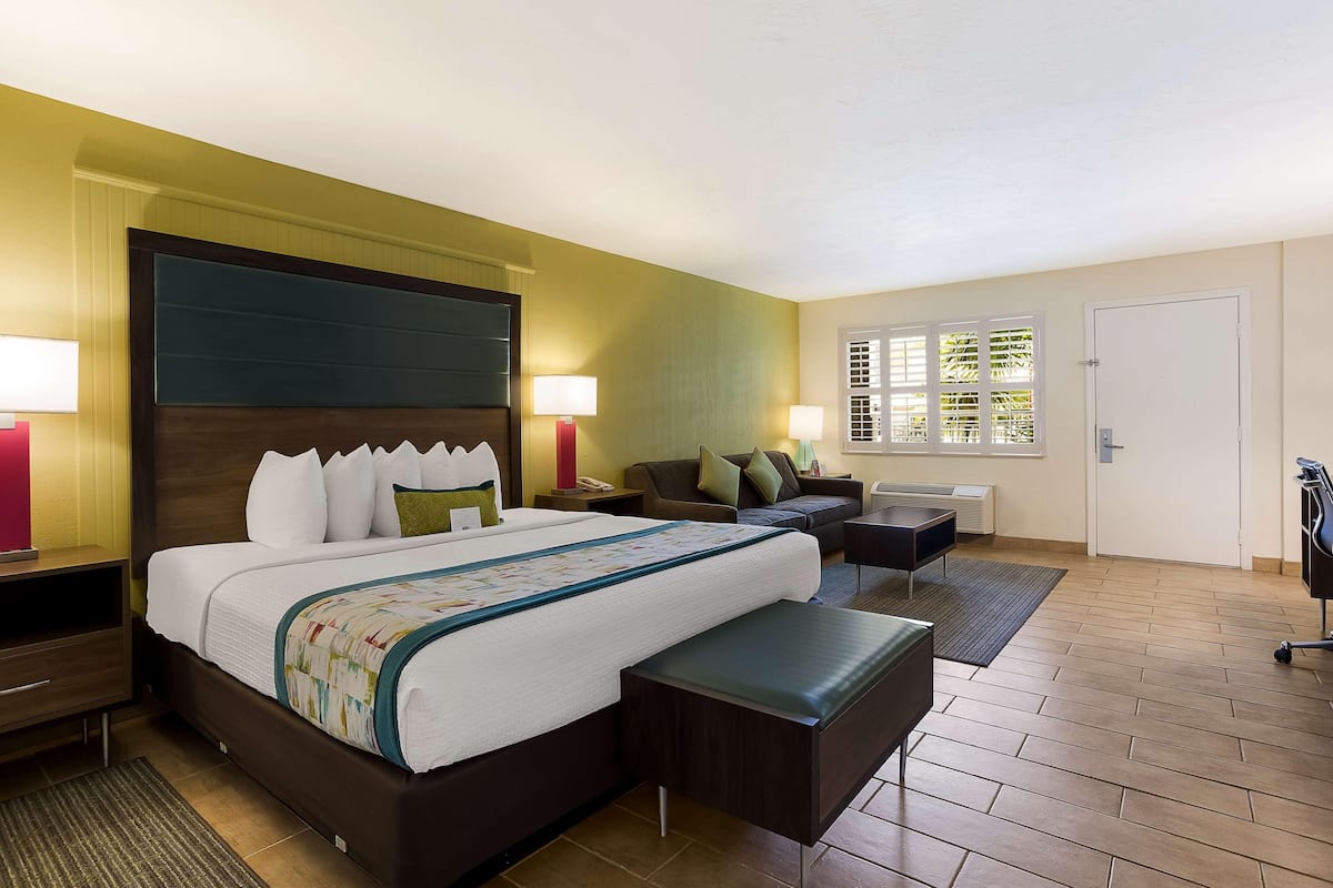 A hotel room with a large bed, bedside tables, a sofa, a desk, and a window with blinds.
