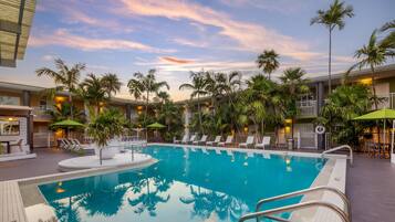 A swimming pool with lounge chairs and umbrellas, surrounded by palm trees and hotel buildings.