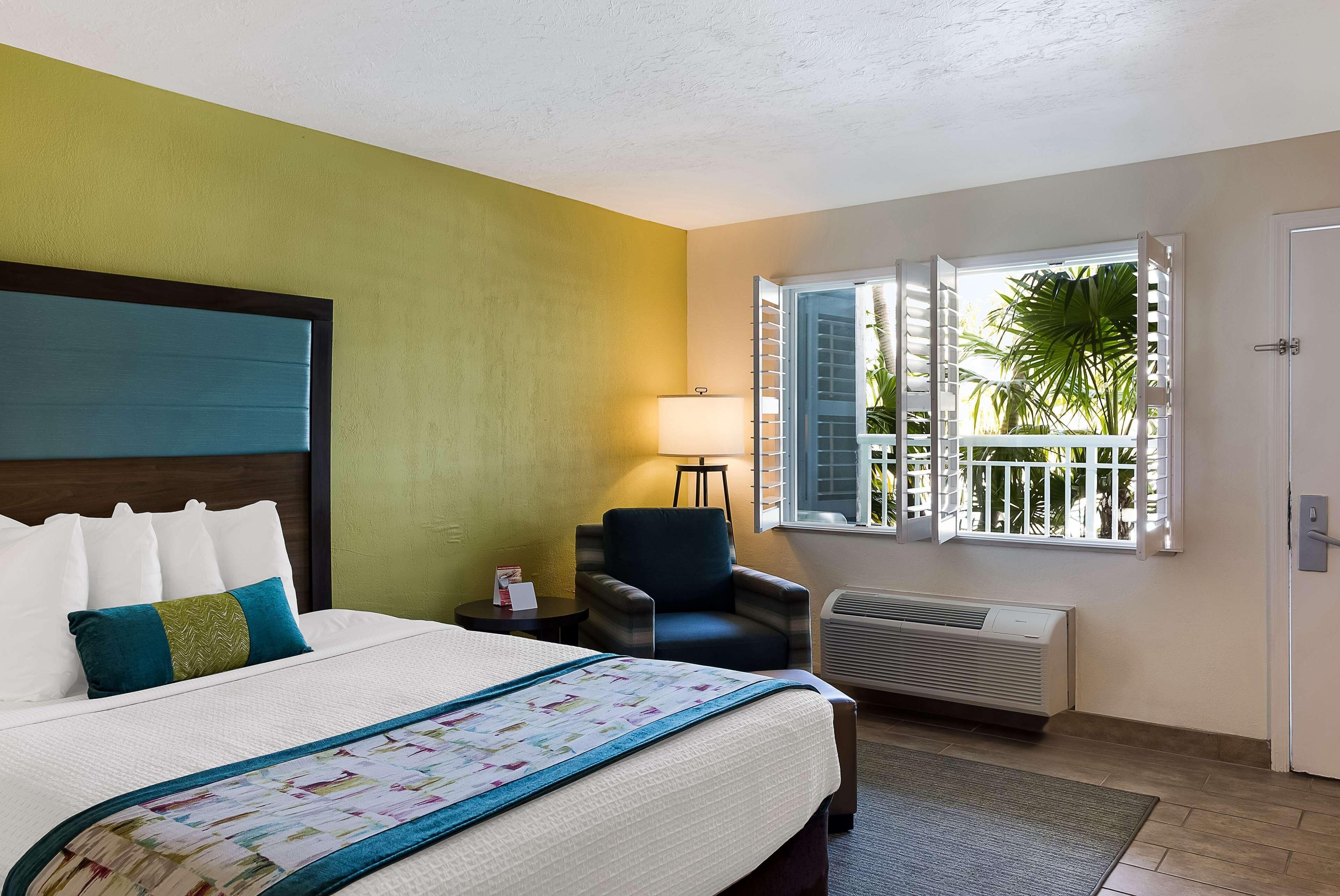 A hotel room with a bed, a chair, a window with a view of palm trees, and an air conditioning unit.