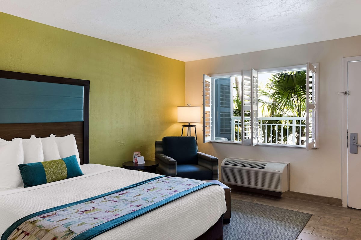 A hotel room with a bed, a chair, a window with a view of palm trees, and an air conditioning unit.